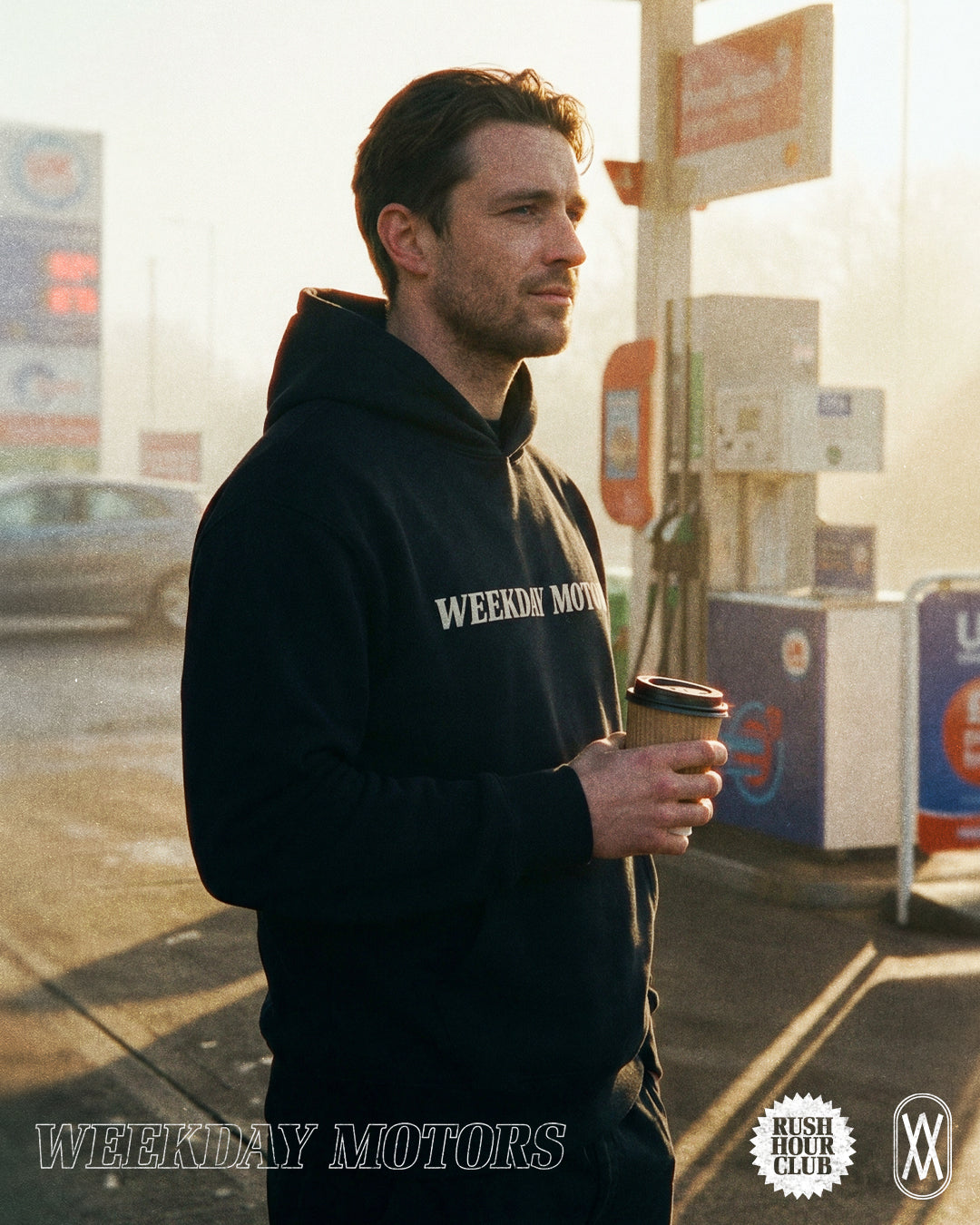 A man wears a black Weeday Motors brand hoodie, drinking a coffee in the early morning light on a petrol station forecourt.