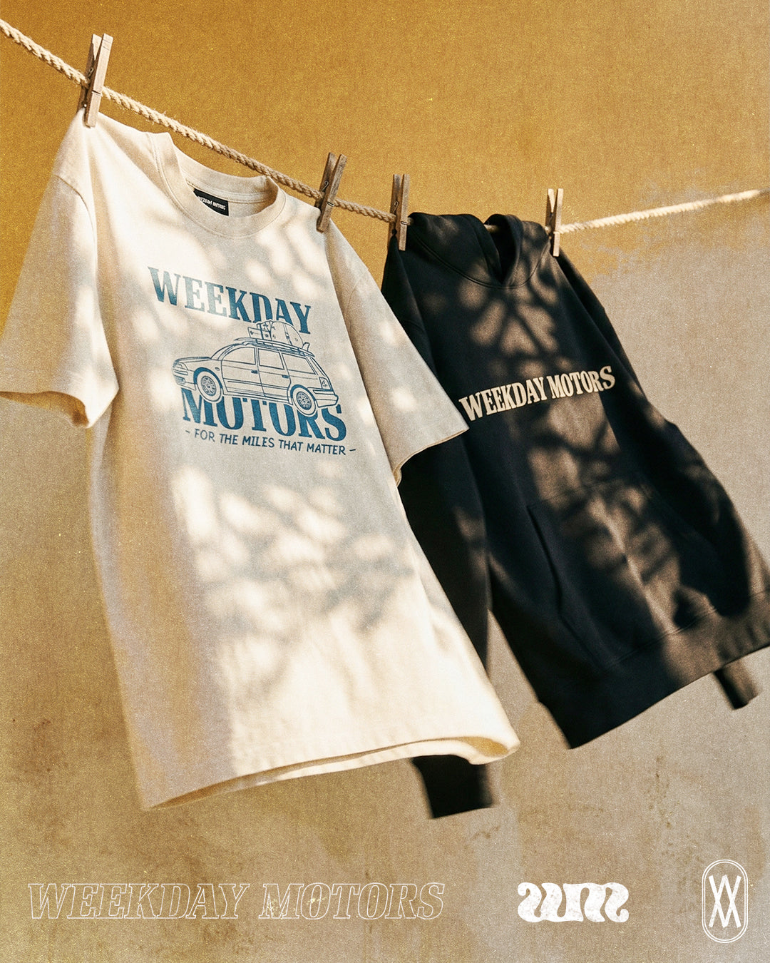 Weekday Motors brand graphic white t-shirt and a black hoodie hang from a washing line, dappled light and a plaster wall in the background.